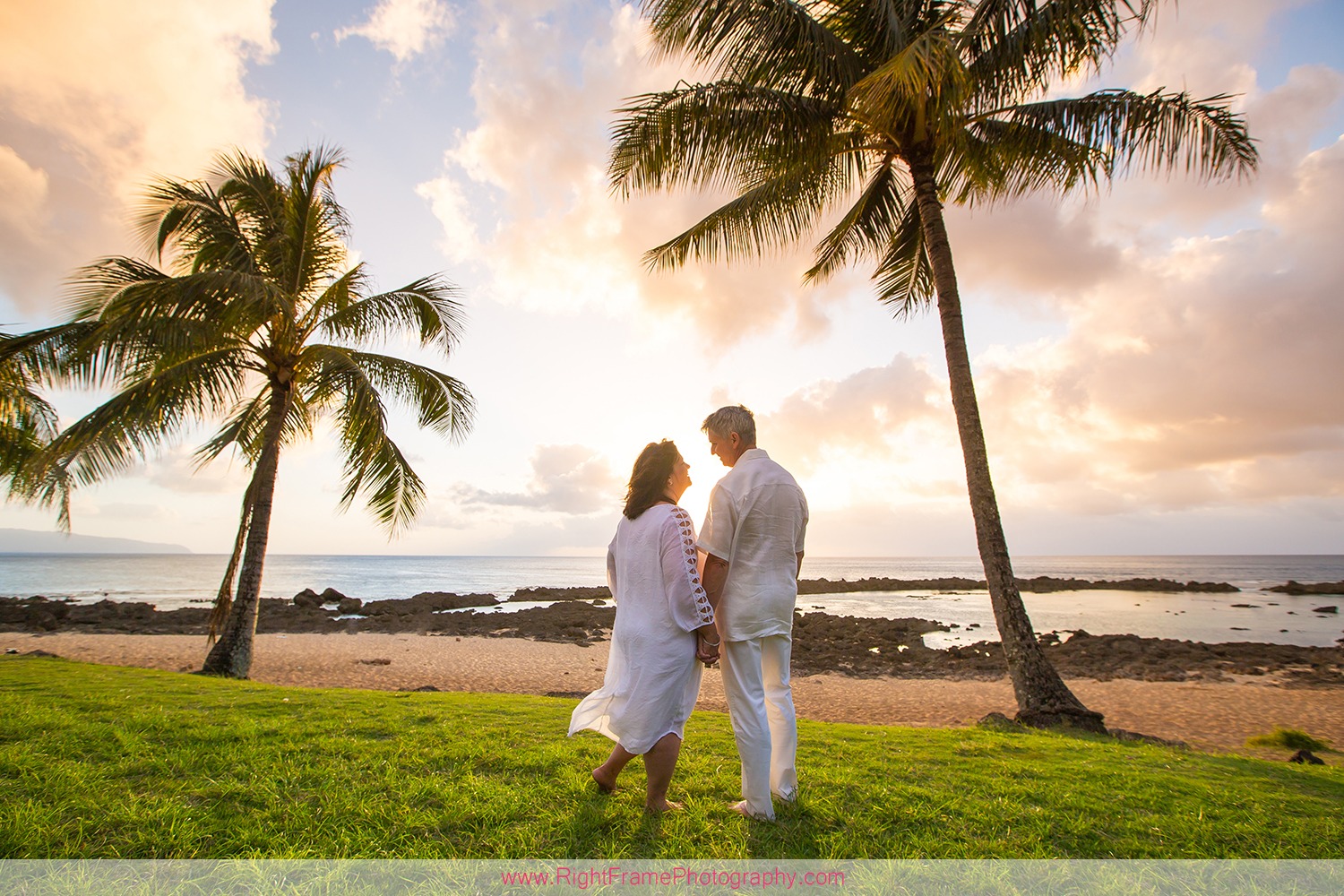 A mature couple in white stands hand-in-hand on a grassy slope overlooking a tropical beach at sunset, framed by palm trees.