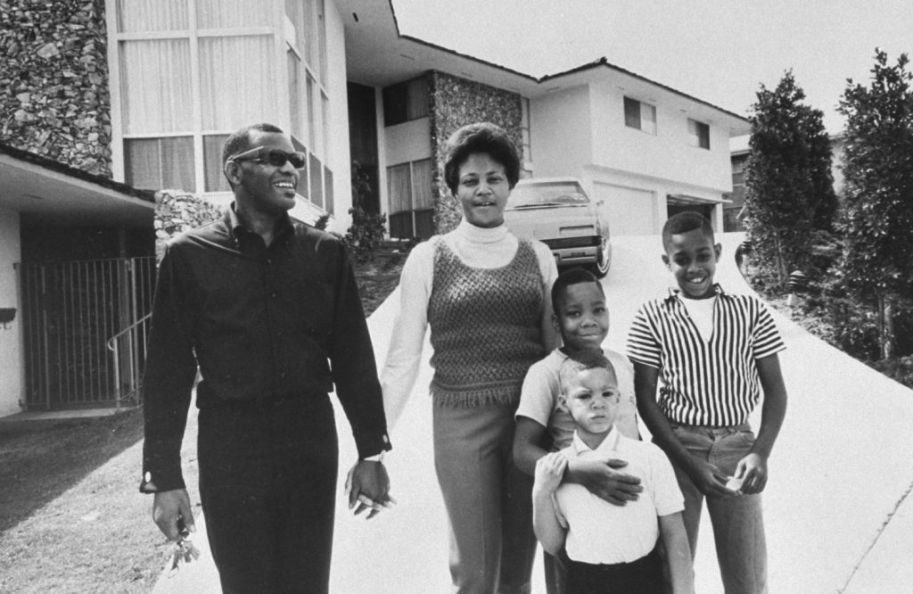 A Black family of five poses smiling on a driveway in front of a mid-century modern home, circa 1960s-70s.