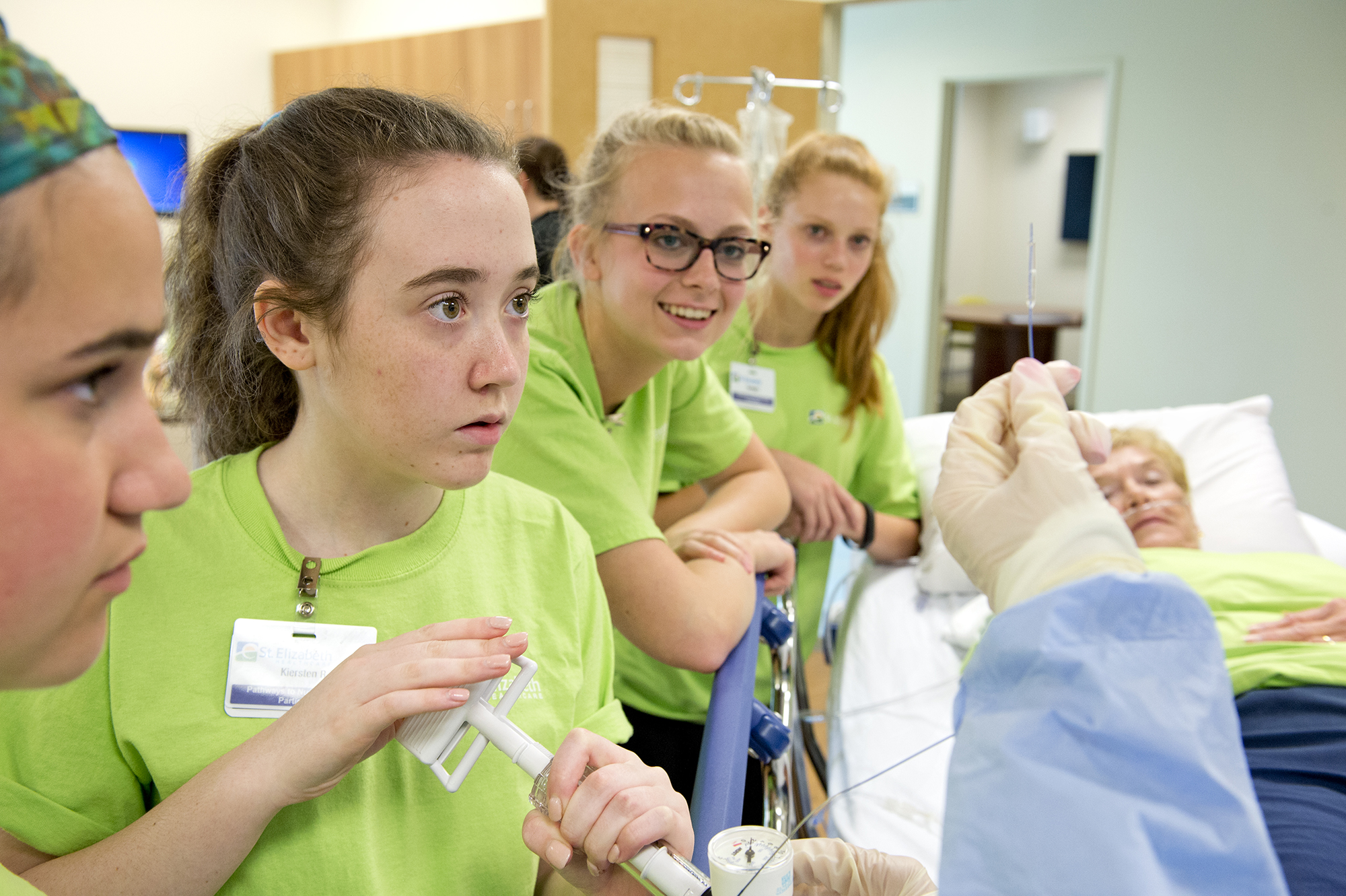 Teenage students in green scrubs observe a medical procedure on a patient in a hospital bed, guided by a clinician.