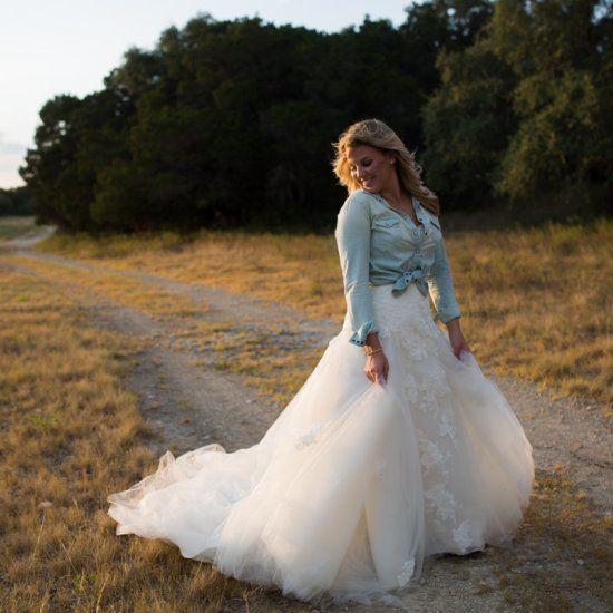 Bride in lace wedding gown with denim jacket, standing on rural dirt path at golden hour.