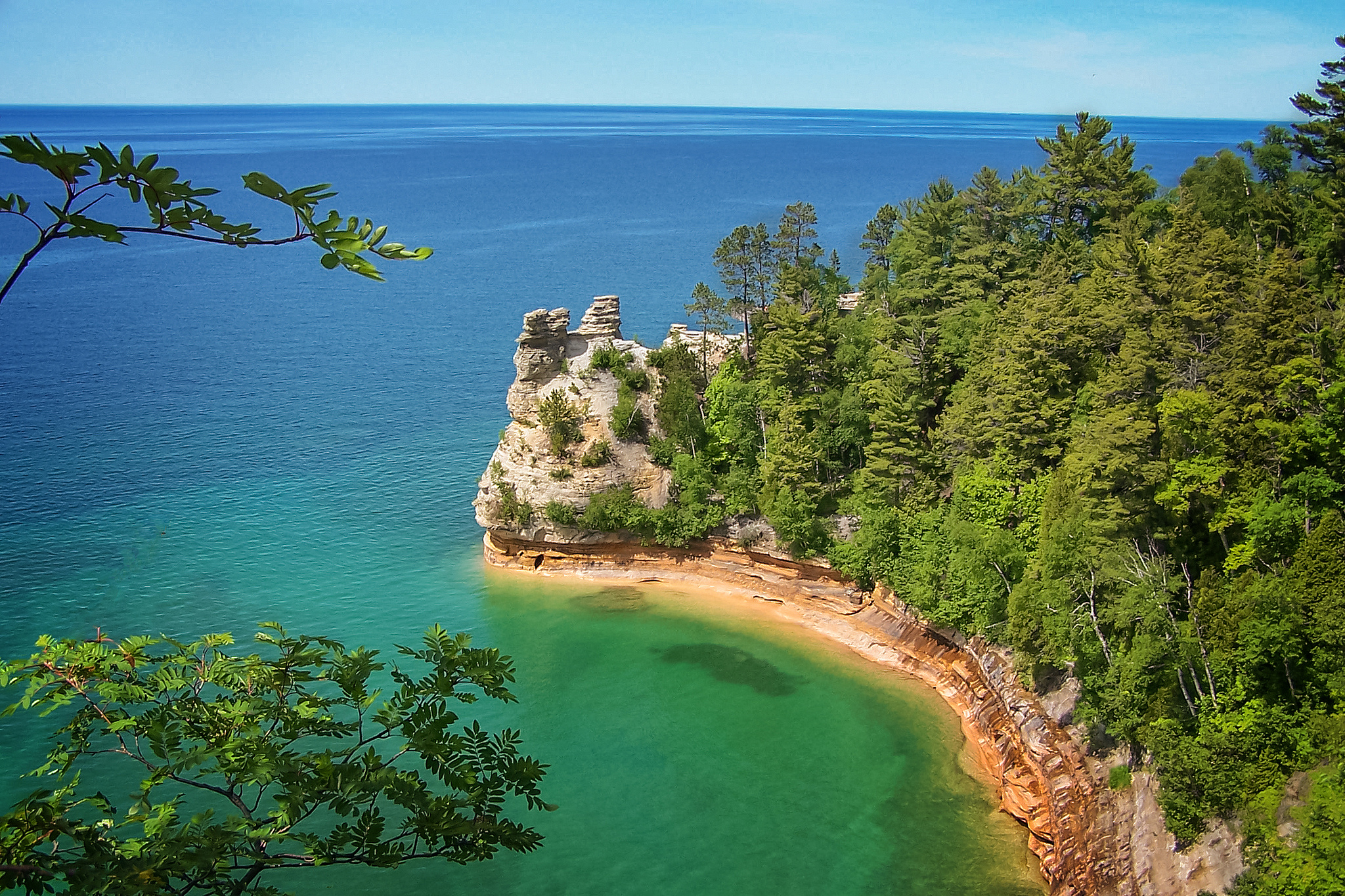 Turquoise waters lap against a rugged sandstone cliff crowned with evergreen forest under a clear blue sky.