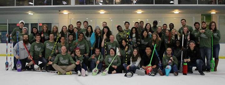 Group of people in matching green shirts posing on an indoor ice rink with broomball sticks and gear.