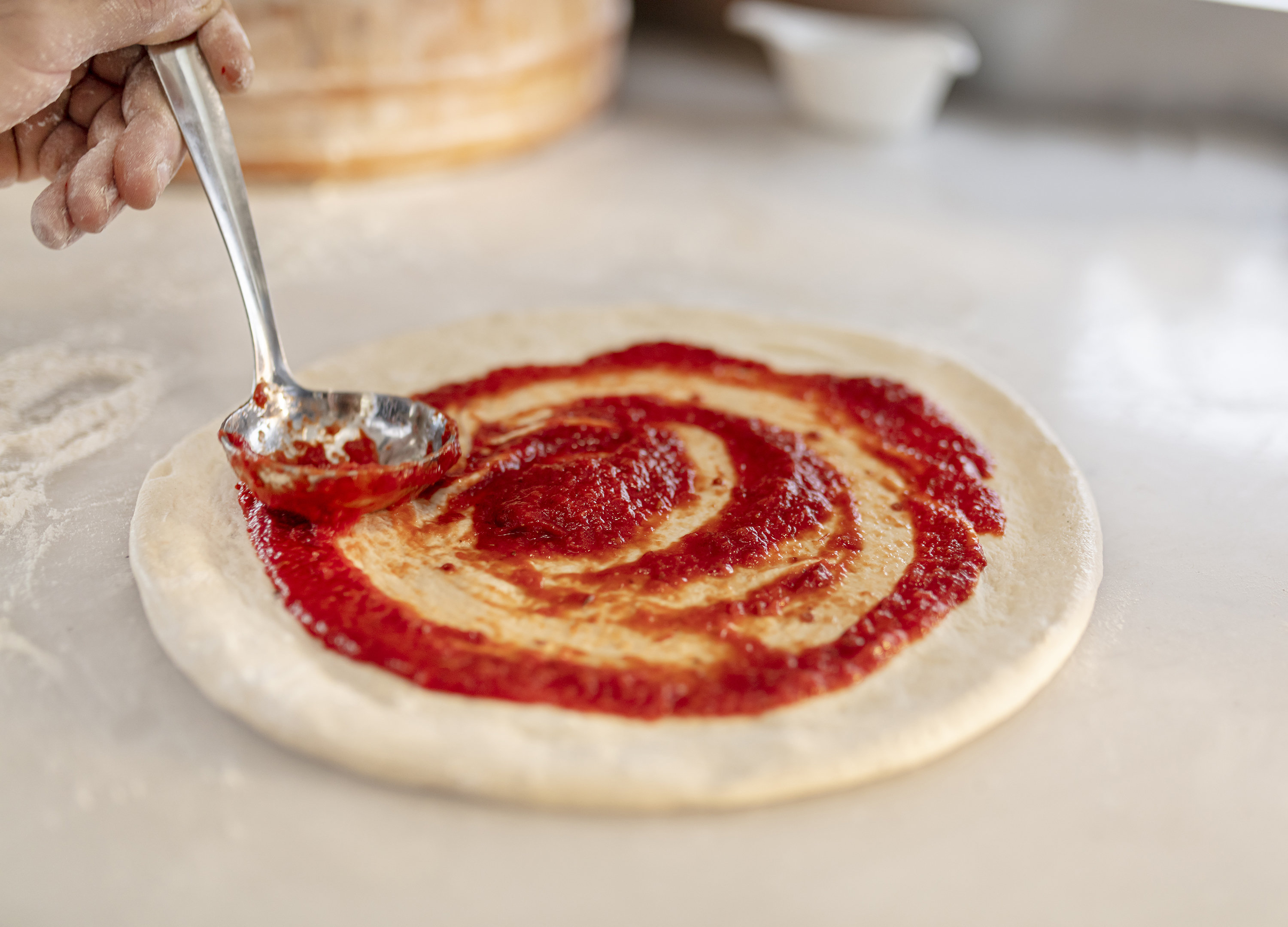 A chef spreads tomato sauce in a spiral on raw pizza dough using a ladle, with flour-dusted hands and a marble countertop.