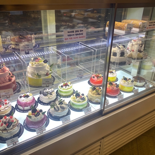 Brightly lit bakery display case filled with colorful fruit-topped cakes and pastries in a Korean café.