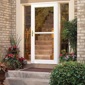 White storm door with screen panel installed on a brick home entrance, flanked by potted flowers and greenery.