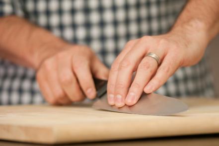 A person in a checkered shirt carefully slices food on a wooden cutting board with a chef’s knife.