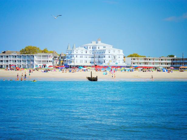 A sunny beach scene with a grand white hotel, colorful umbrellas, swimmers, and a small boat near shore under a clear blue sky.