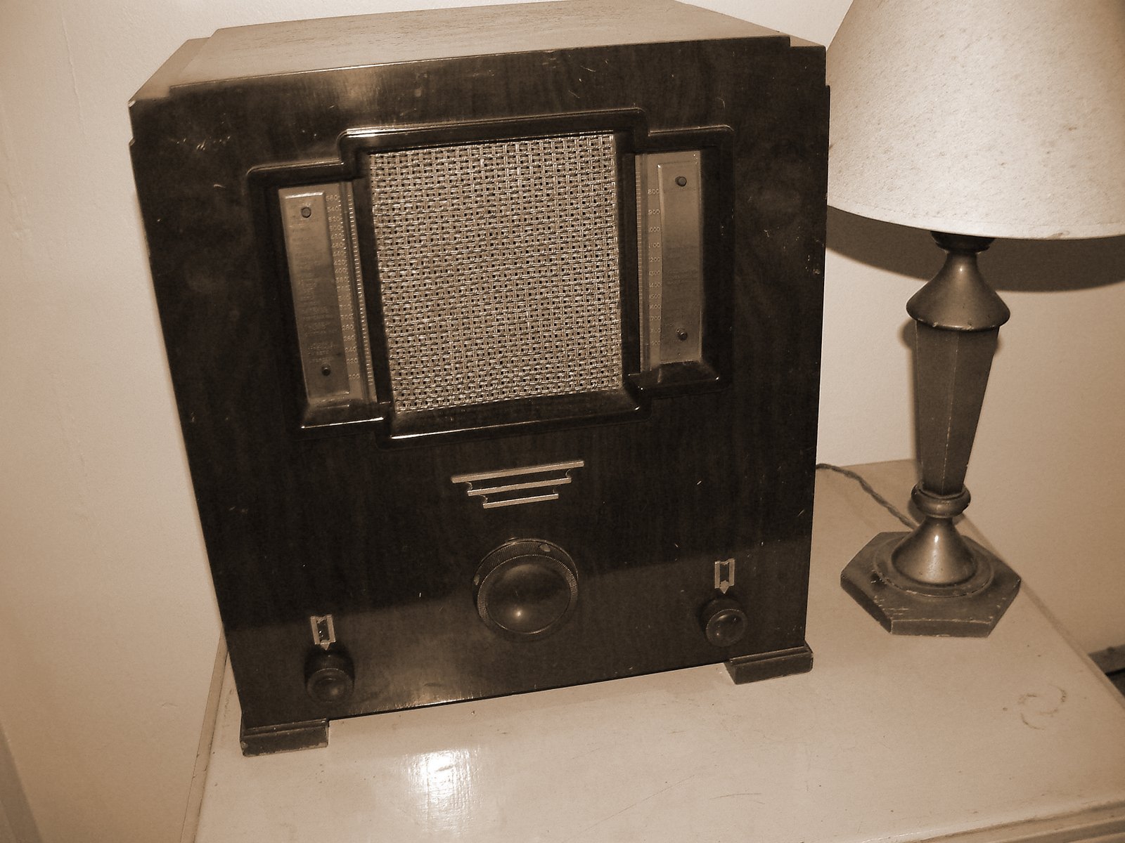 Vintage wooden radio with metal grille, tuning dials, and knobs beside a classic table lamp on a nightstand.