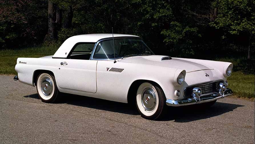 White 1956 Ford Thunderbird convertible parked on asphalt with trees in background.
