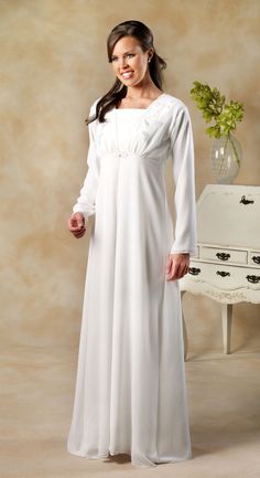 Woman wearing a white modest long-sleeve floor-length dress with square neckline and subtle waist detail, standing beside a vintage dresser.