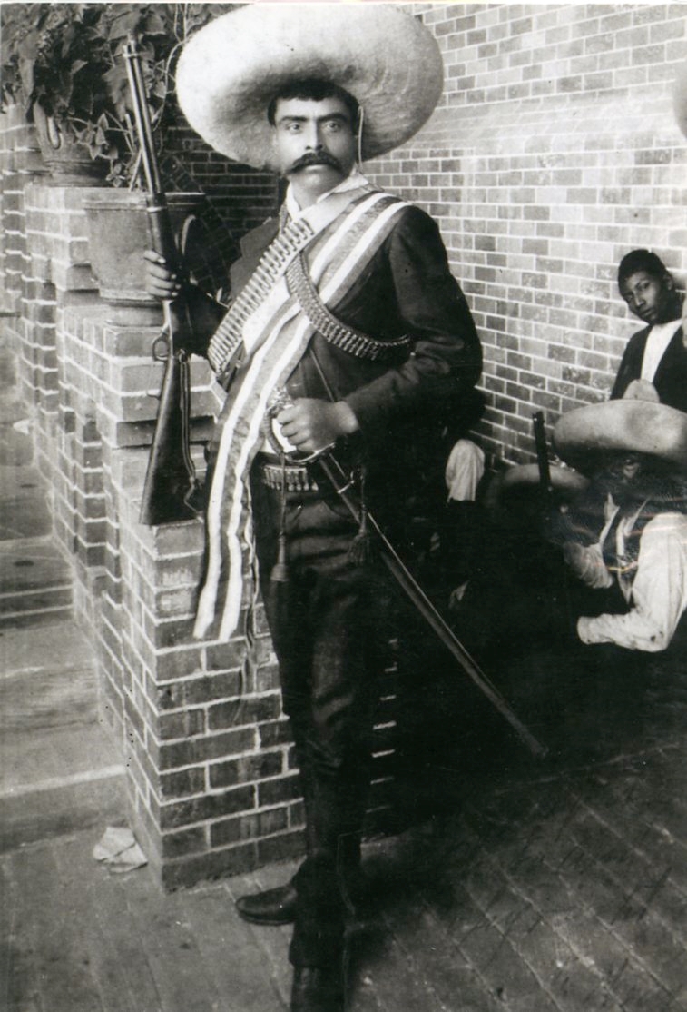 A mustachioed man in a large sombrero, sash, and bandolier holds a rifle beside a brick wall; others sit nearby.