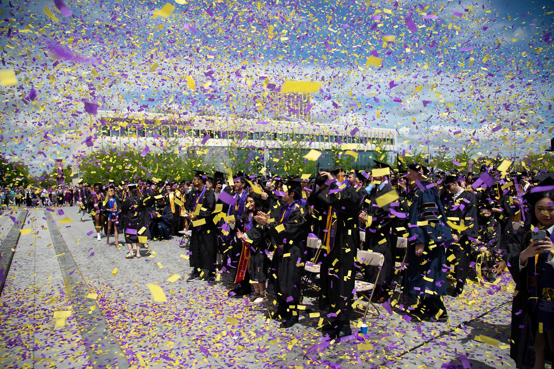 Graduates in black gowns and purple-yellow stoles celebrate under a shower of confetti at an outdoor commencement ceremony.