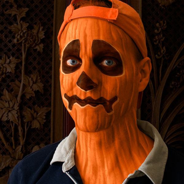 Person wearing a realistic pumpkin mask with carved facial features and an orange cap, set against ornate wooden paneling.