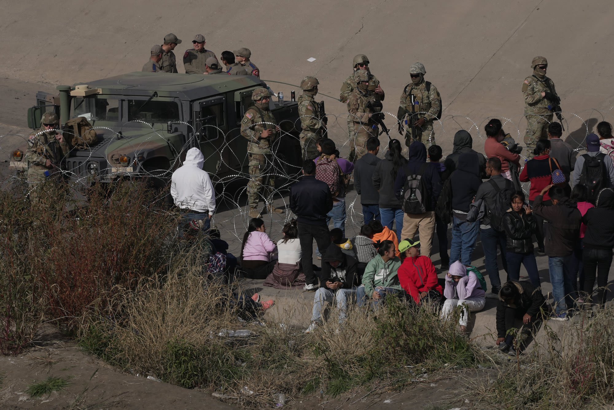 U.S. military personnel and border agents stand near barbed wire as migrants wait on the ground near a Humvee at the U.S.-Mexico border.