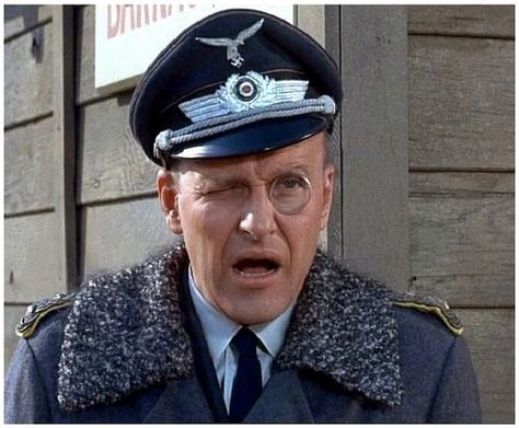 German Luftwaffe officer in WWII-era uniform with peaked cap, round glasses, and fur-collared coat, expressing surprise or alarm.