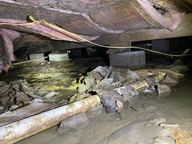 Severely flooded crawl space with muddy water, damaged insulation, rusted pipes, and debris under a home’s foundation.