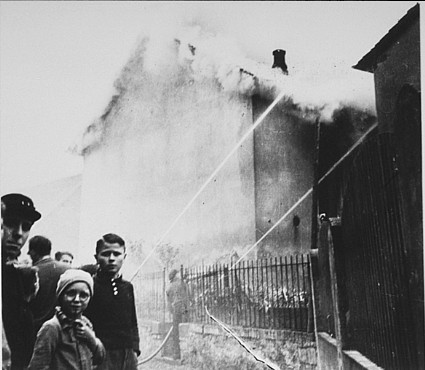 Firefighters spray water on a burning building as onlookers, including children, watch in concern.