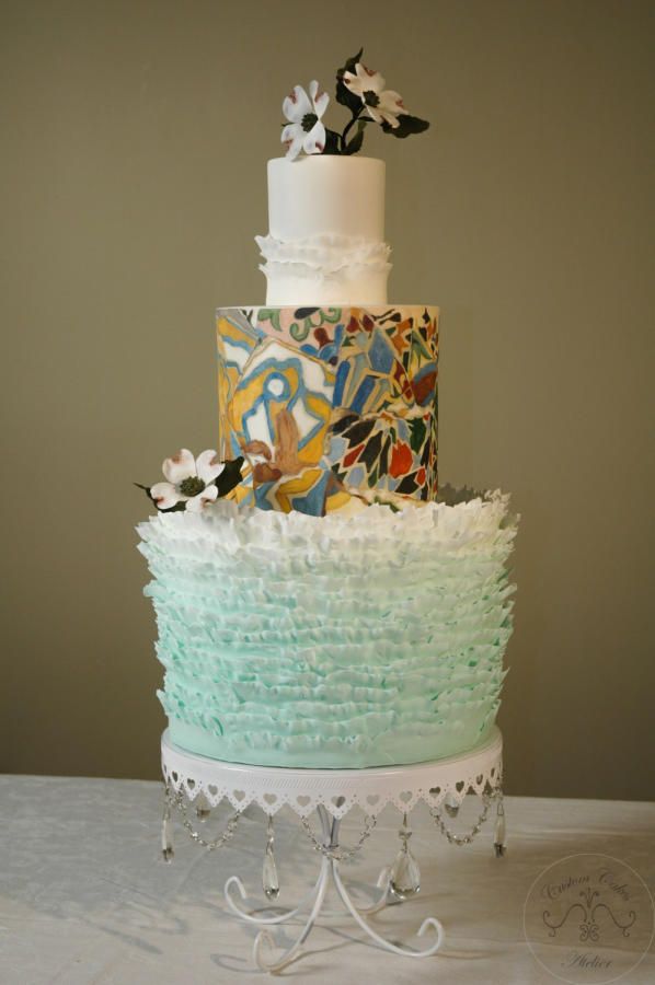 Three-tiered artistic wedding cake with ruffled mint base, mosaic middle, and floral topper on ornate stand.