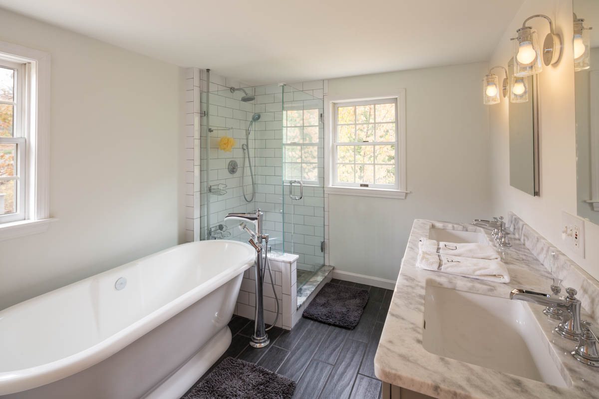 Bright, modern bathroom with freestanding tub, glass-enclosed shower, marble double vanity, and natural light from two windows.