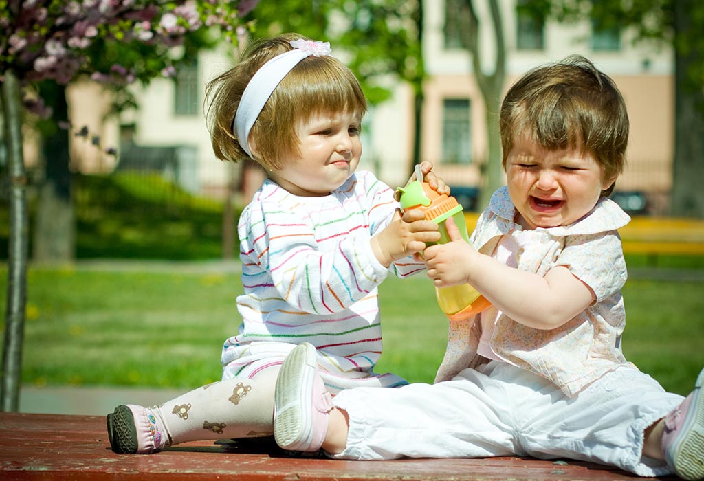 Two toddlers sitting on a bench outdoors, one smiling while the other cries, both tugging at a sippy cup.