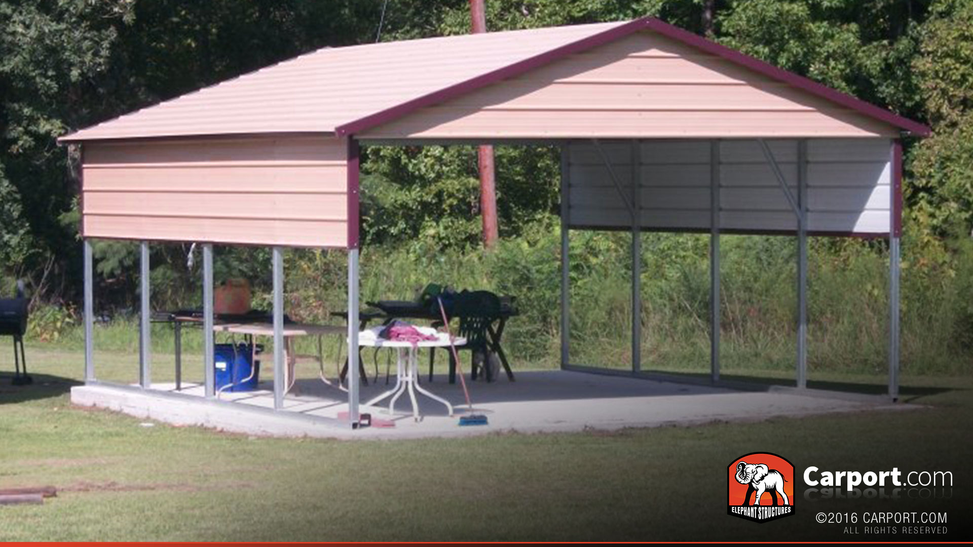 Beige metal carport with maroon trim, open sides, concrete slab base, and outdoor furniture underneath in a grassy yard.