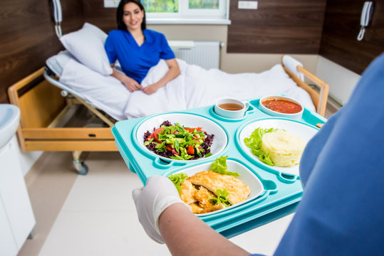 A healthcare worker in blue scrubs serves a balanced meal tray to a smiling patient in a hospital bed.