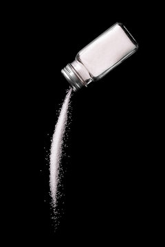 Glass salt shaker倾倒 white granular salt against black background, high-contrast studio shot.
