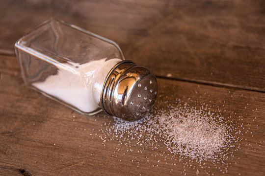 A clear glass salt shaker lying on its side on a wooden surface, spilling fine white salt crystals.