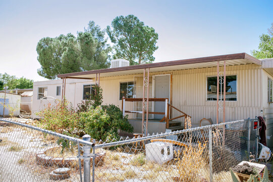 Weathered single-wide mobile home with porch, chain-link fence, and overgrown yard under clear sky.