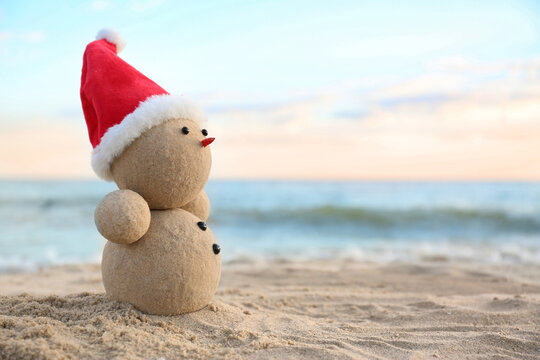 A sand snowman wearing a red Santa hat on a beach at sunset, with ocean waves in the background.