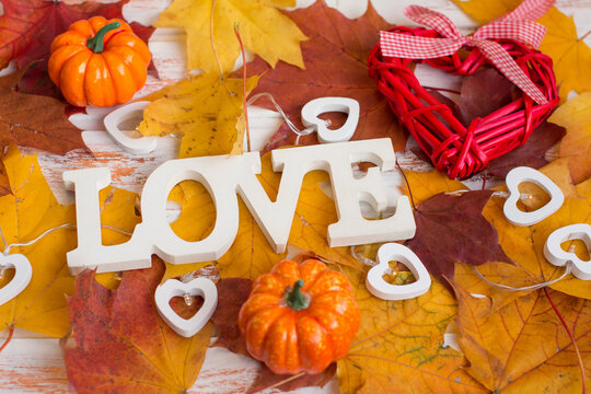 Autumn-themed "LOVE" sign surrounded by fall leaves, mini pumpkins, and heart decorations on a rustic wooden background.
