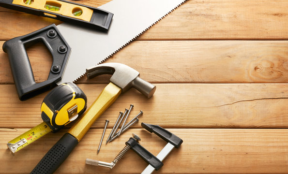 A variety of hand tools including a saw, hammer, tape measure, level, clamp, and nails arranged on a wooden workbench.