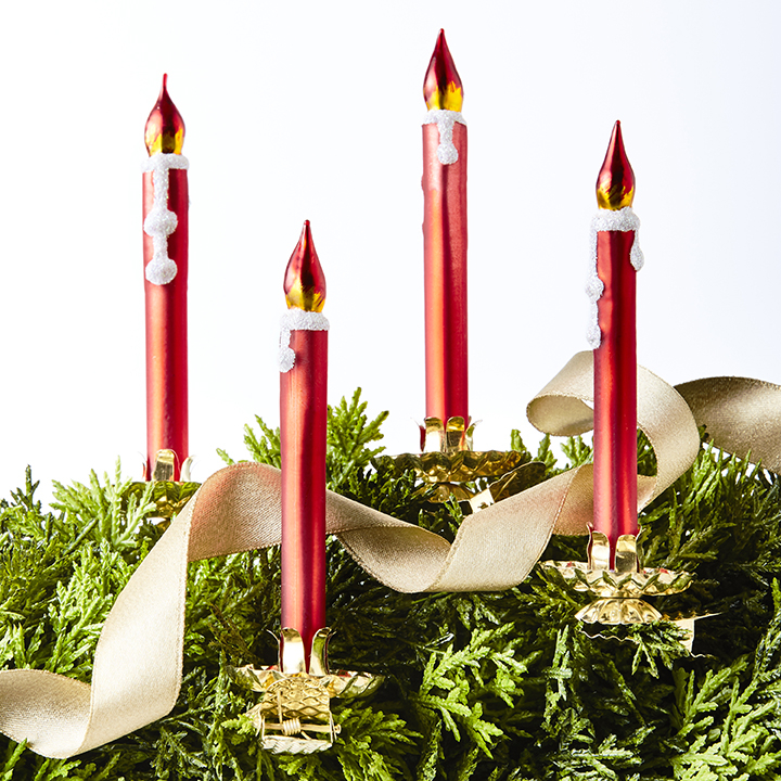 Four red Advent candles with gold flame tips and white wax drips, arranged on greenery with a cream ribbon and gold holders.