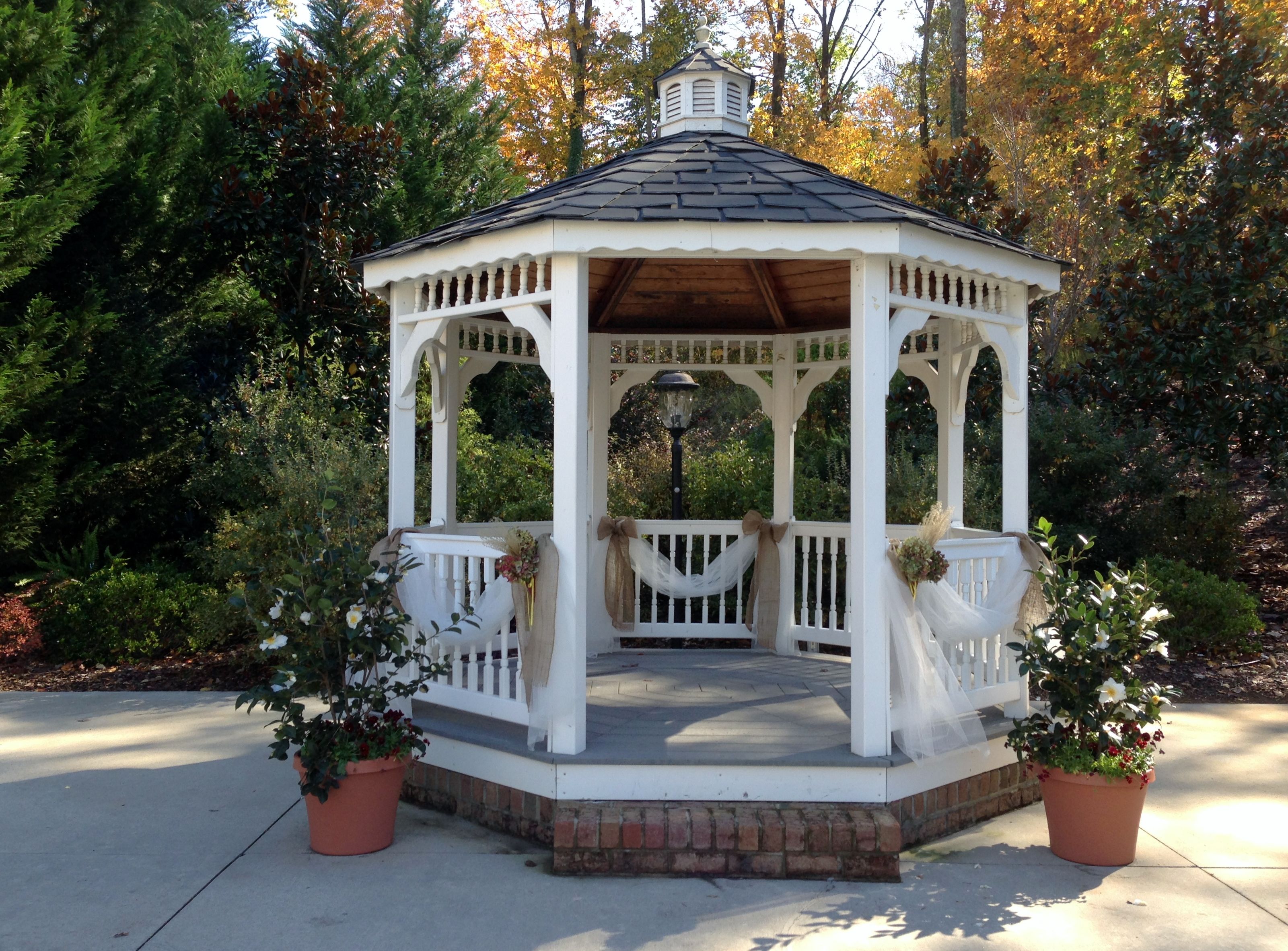 White octagonal gazebo with black shingled roof, decorated with burlap, white fabric, and floral arrangements, set in autumn garden.