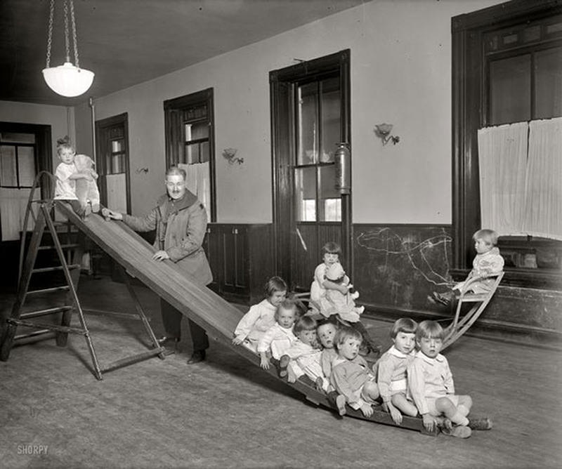Early 20th-century indoor playground scene with children on a wooden slide supervised by an adult man.