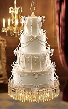 Ornate multi-tiered white wedding cake with pearl drapery, crystal chandelier base, and hanging from a gold chain in opulent setting.