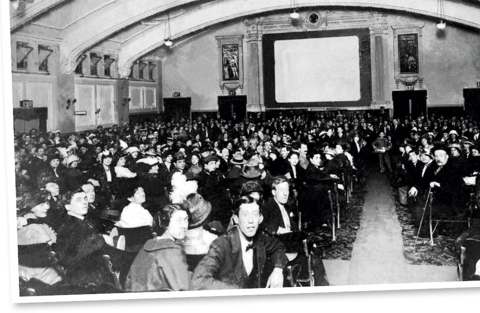 Crowded early 20th-century movie theater with ornate decor, audience in period attire watching a silent film screening.