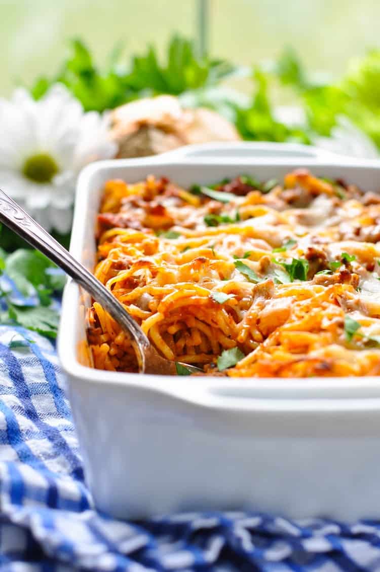 Baked spaghetti casserole with melted cheese, herbs, and a serving spoon in a white dish on a blue checkered cloth.