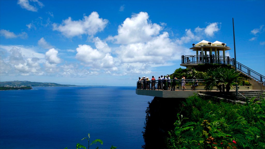 Tourists admire the vast blue ocean from a cliffside viewing platform under a bright sky with scattered clouds.