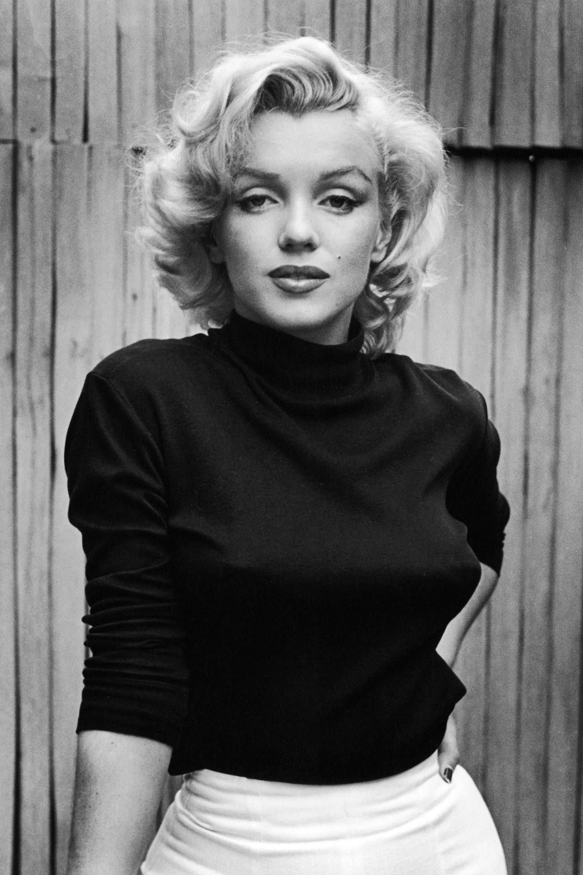 Marilyn Monroe in a black turtleneck and white skirt, posing against a wooden fence, 1950s black-and-white portrait.