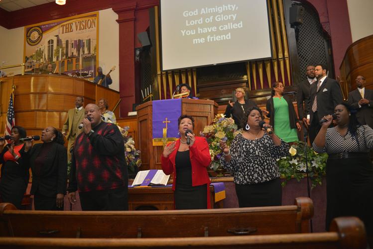 A diverse gospel choir sings passionately in a church sanctuary with a projection screen displaying worship lyrics.