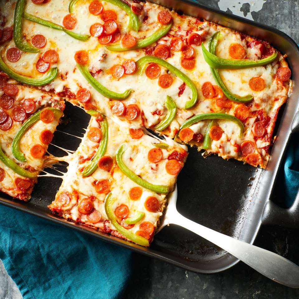 Baked pepperoni and green bell pepper pizza squares in a metal pan, one slice lifted with a spatula showing gooey cheese.