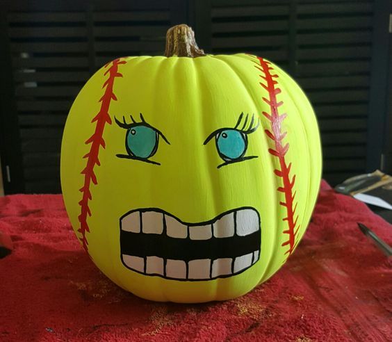 Yellow softball-painted pumpkin with expressive eyes, red stitching, and a grinning mouth on a red cloth background.