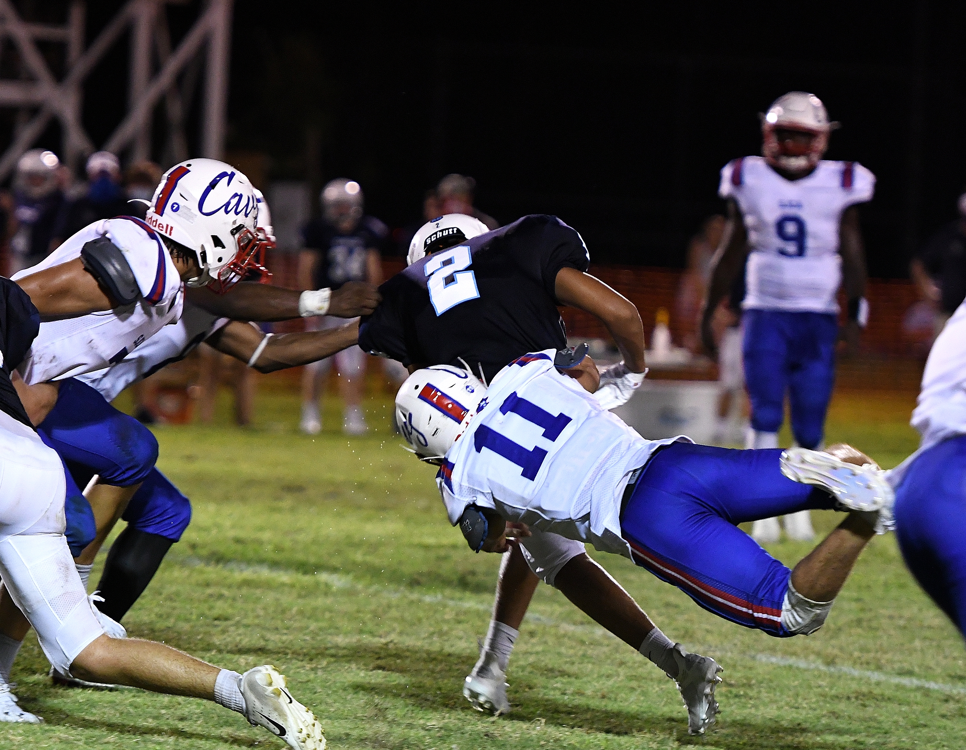 High school football player in white and blue uniform (No. 11) dives to tackle opponent in black jersey (No. 2) during night game.