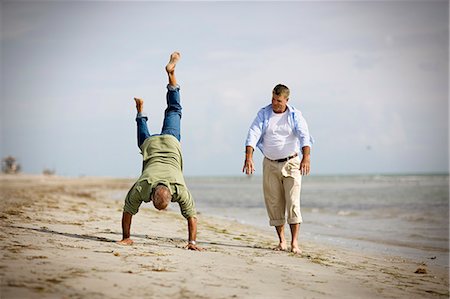 Two men on a beach-one doing a handstand, the other walking nearby-capturing joy and balance in a casual seaside moment.