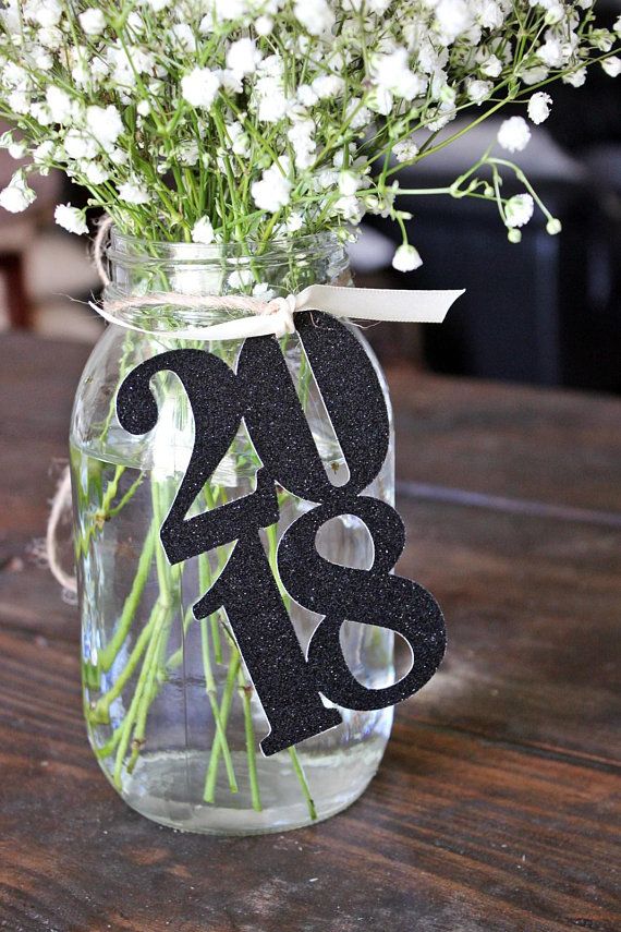 Mason jar with baby’s breath and glittery "2018" cutout, tied with ribbon on wooden table.