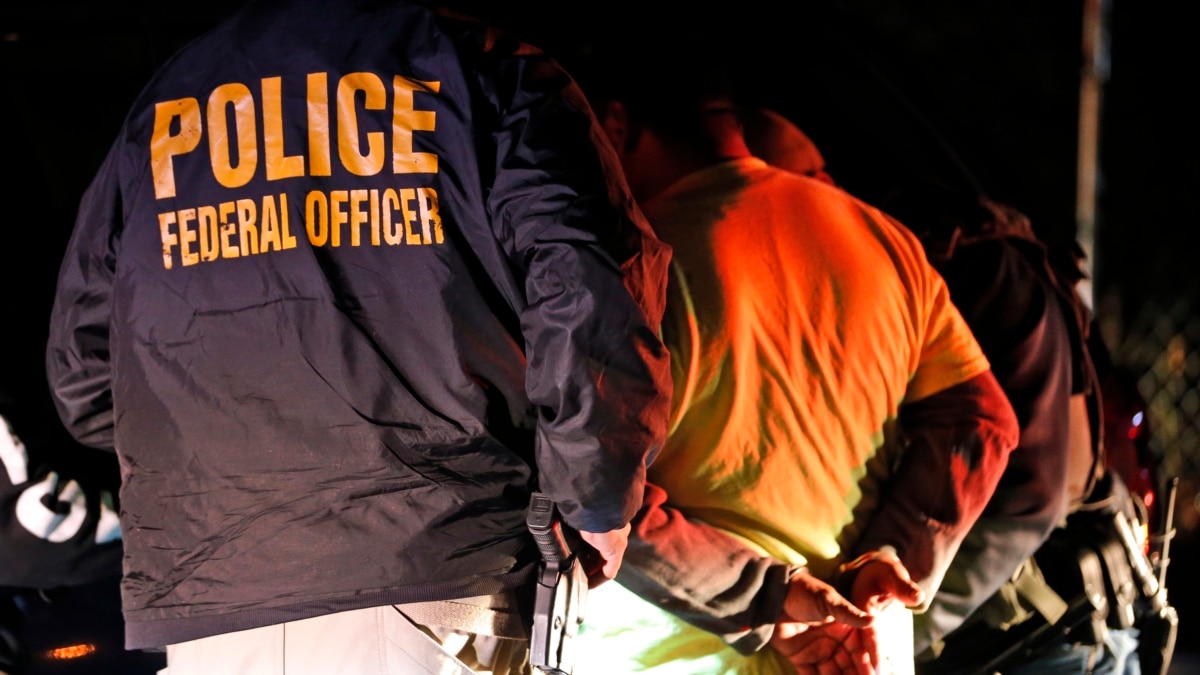 Federal officer in navy jacket with "POLICE FEDERAL OFFICER" text, overseeing a nighttime arrest of individuals in handcuffs.