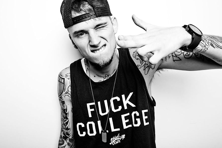Tattooed young man in backward cap and "FUCK COLLEGE" tank top making a hand gesture and grimacing playfully.
