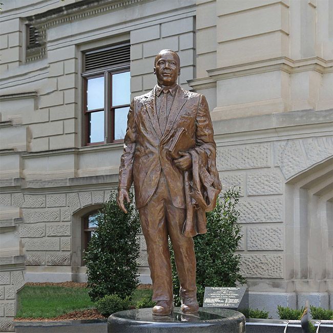 Bronze statue of a man in a suit holding papers, standing before a stone government building.