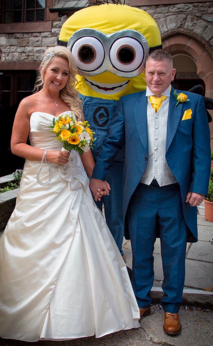 Bride and groom pose with a Minion character at their wedding outside a stone building.